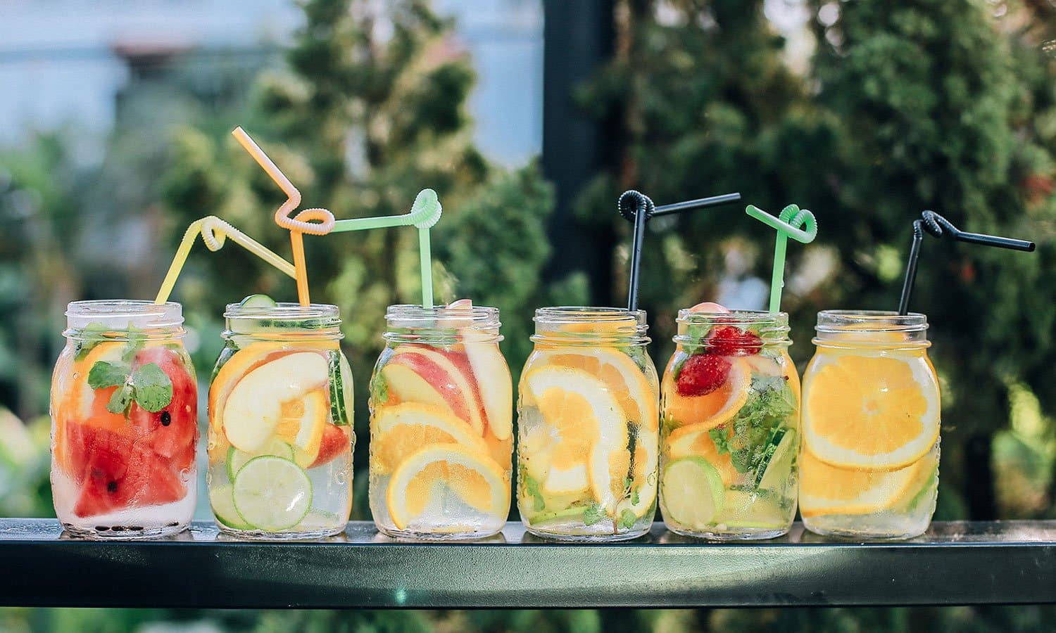 Six fruit-infused jars of water with straws on an outdoor table.